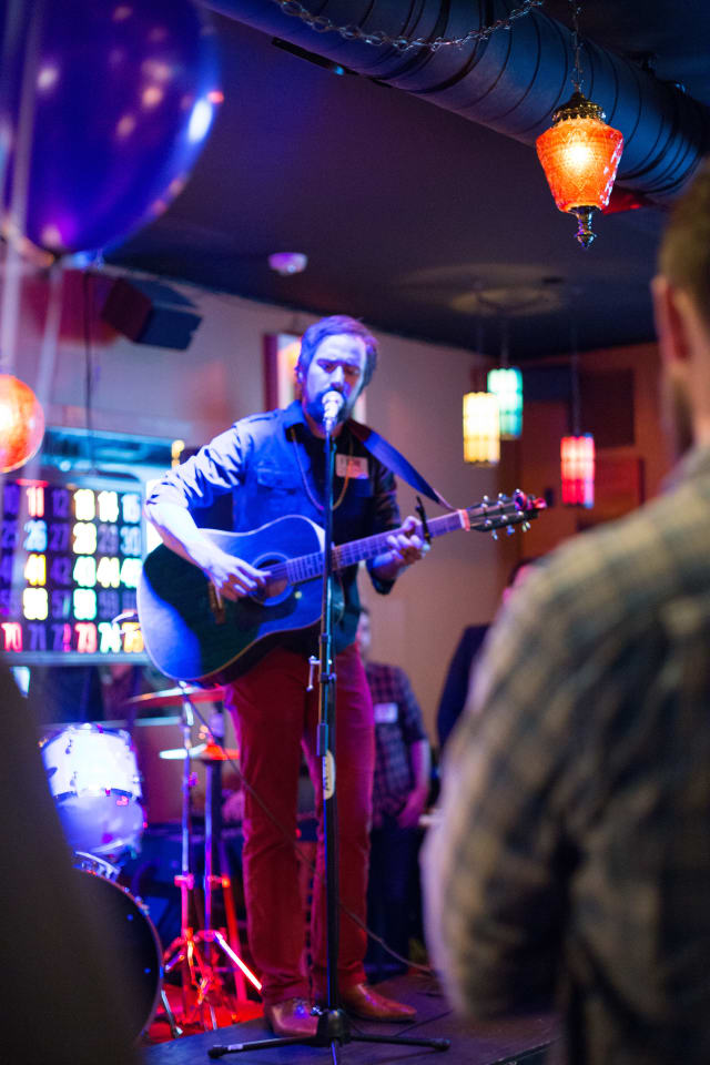 Musician performing with an acoustic guitar on stage at the Love Bomb 2017 event, illuminated by colorful lights, with audience members visible in the foreground.